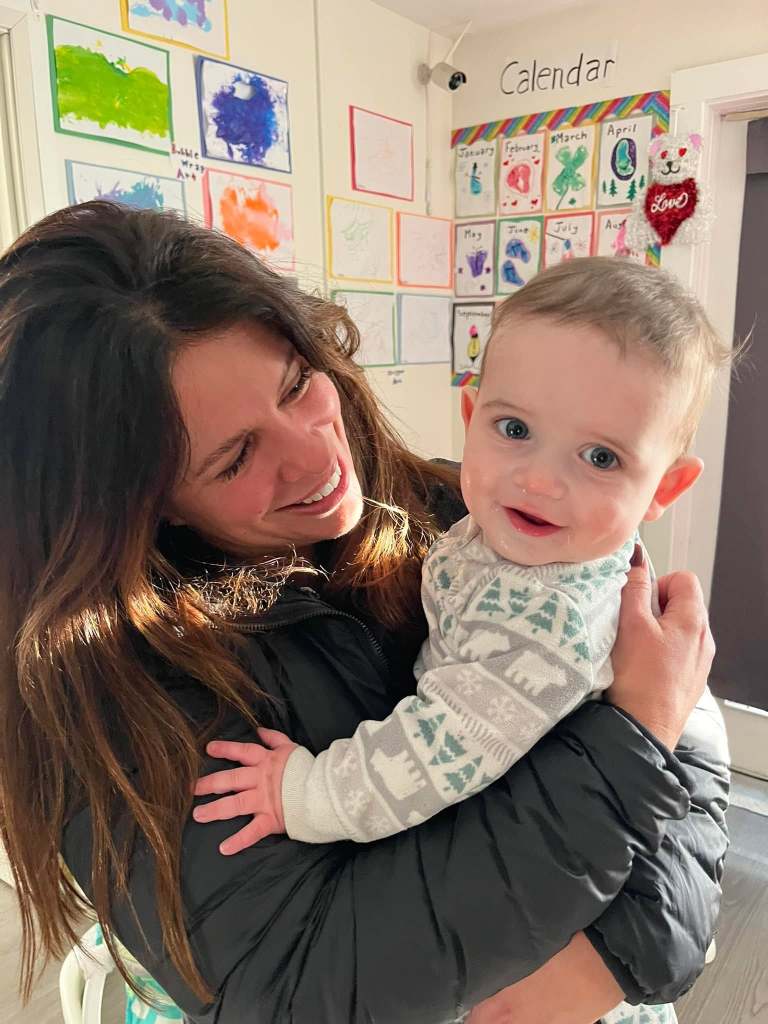 A woman smiles joyfully while holding a happy baby in her arms, with colorful children's artwork displayed on the walls behind them.